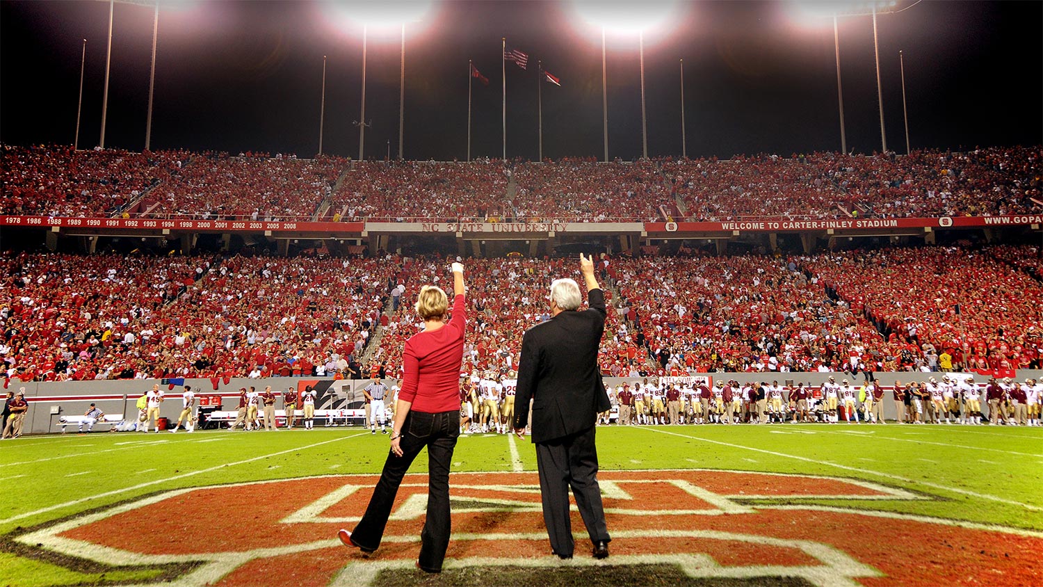 Chancellor Randy Woodson and his wife, Susan, face the crowd while being introduced during a timeout in a football game.