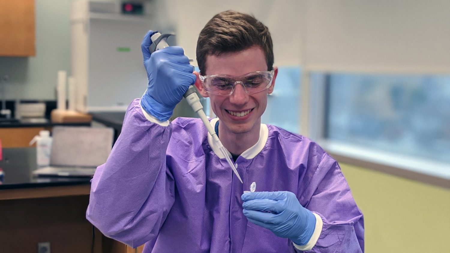 Daniel Heller holds a test tube and pipette in the lab while wearing protective goggles, gloves and a gown.