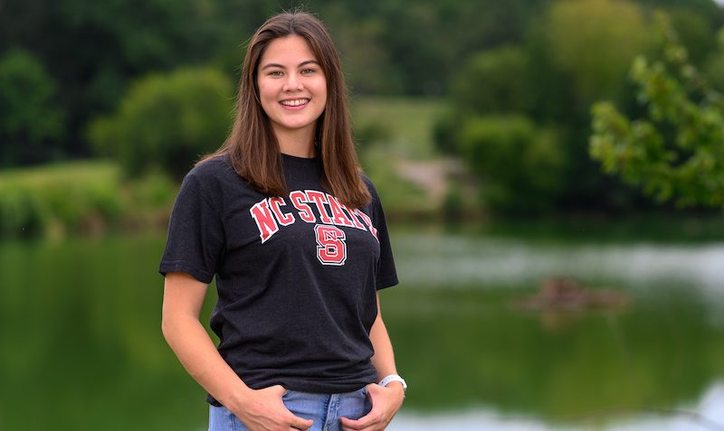 Raena Eldridge stands in front of a pond in an NC State shirt
