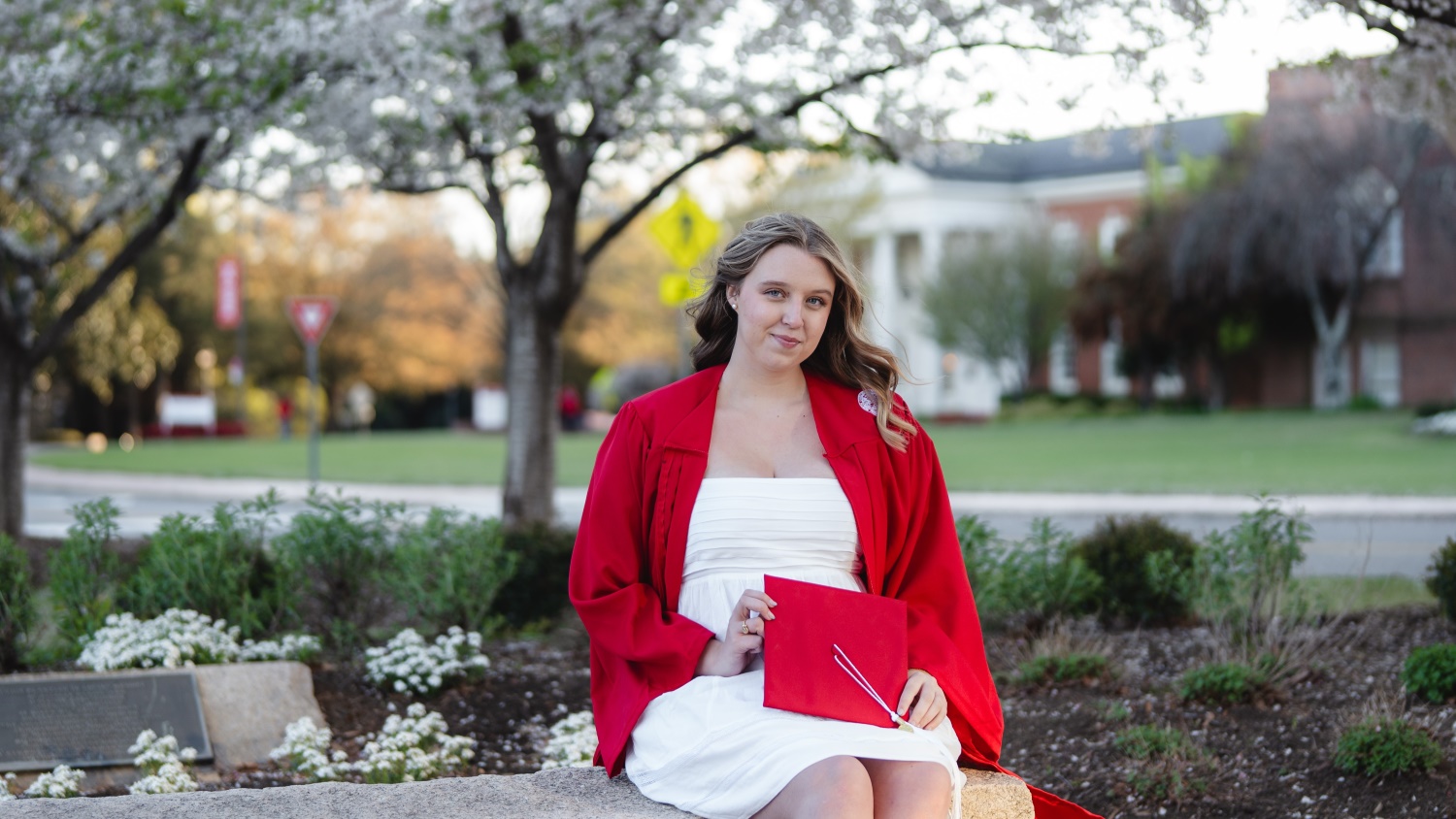 Olivia Ball sitting on a bench among flowering plants in her graduation gown holding her cap.
