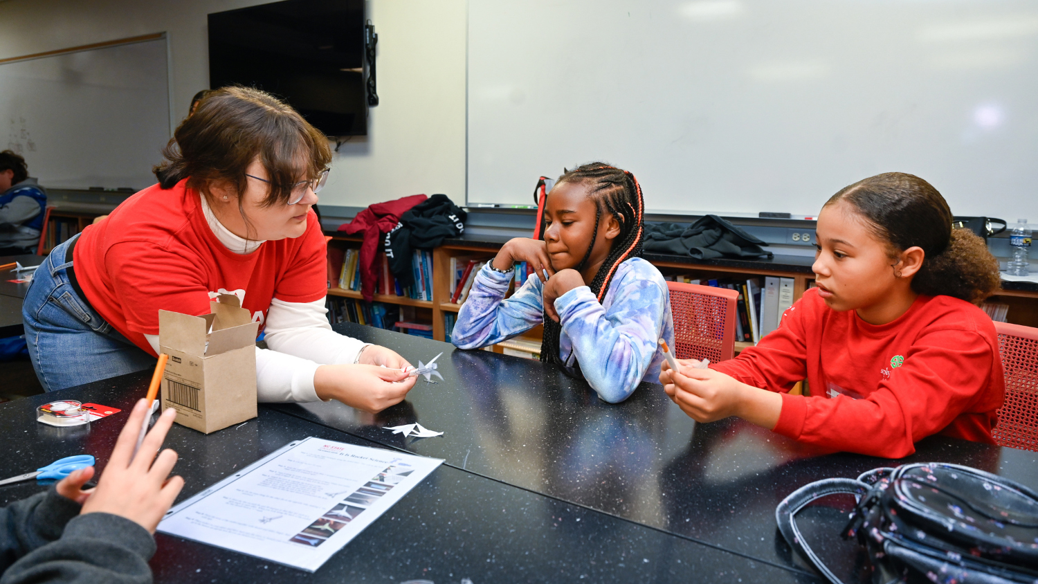 An NC State student, left, teaches two younger girls at a table.