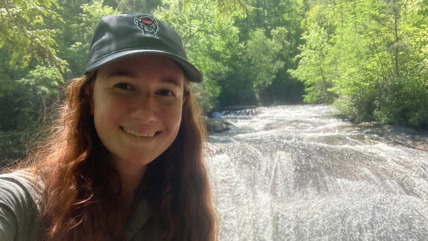 Caitlin McDade wearing an NC State baseball cap in front of a waterfall