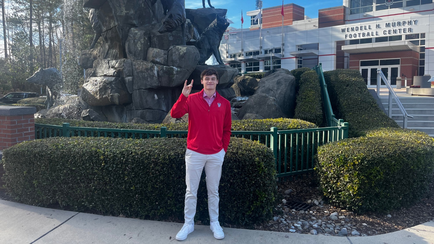 Banks Cathey holding up the wolfie hand sign in front of the wolf statues outside the Wendell H. Murphy Football Center.