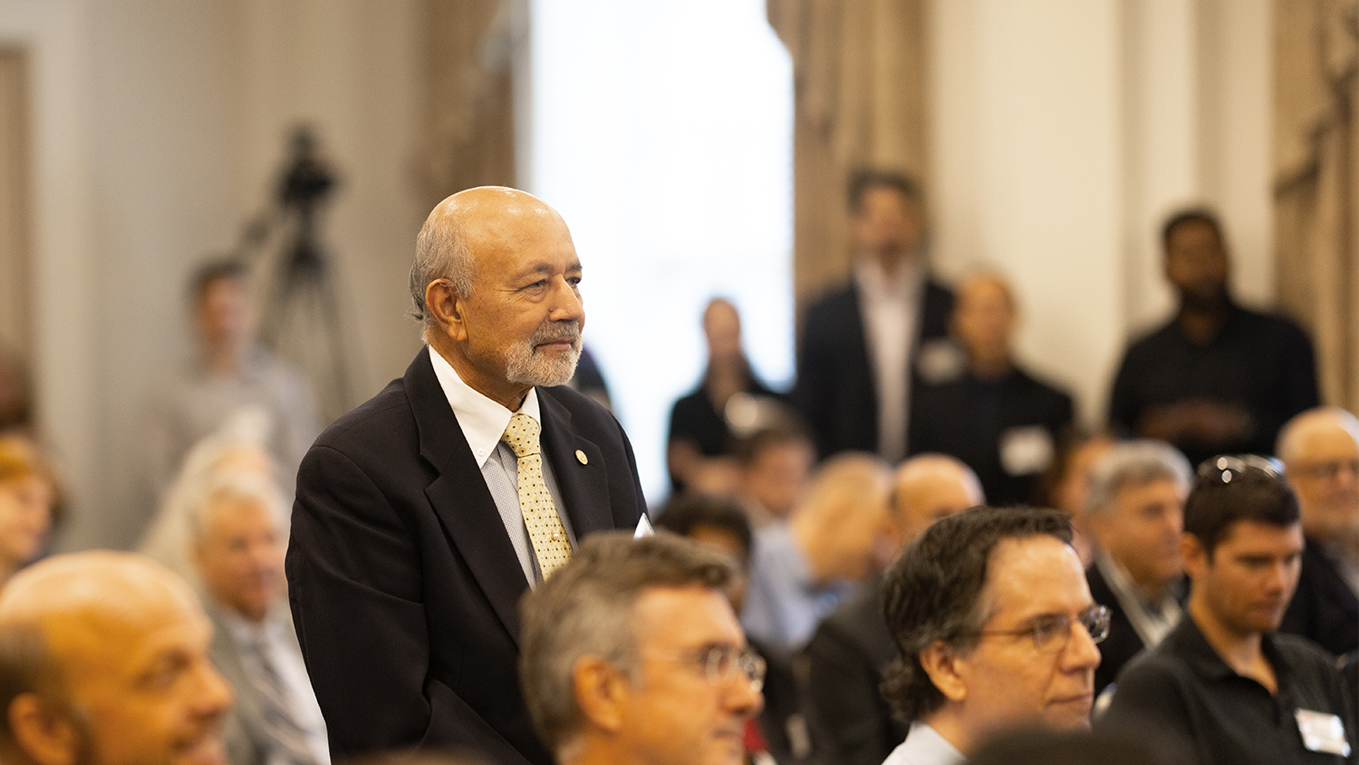 B. Jayant Baliga standing in a room of seated people.