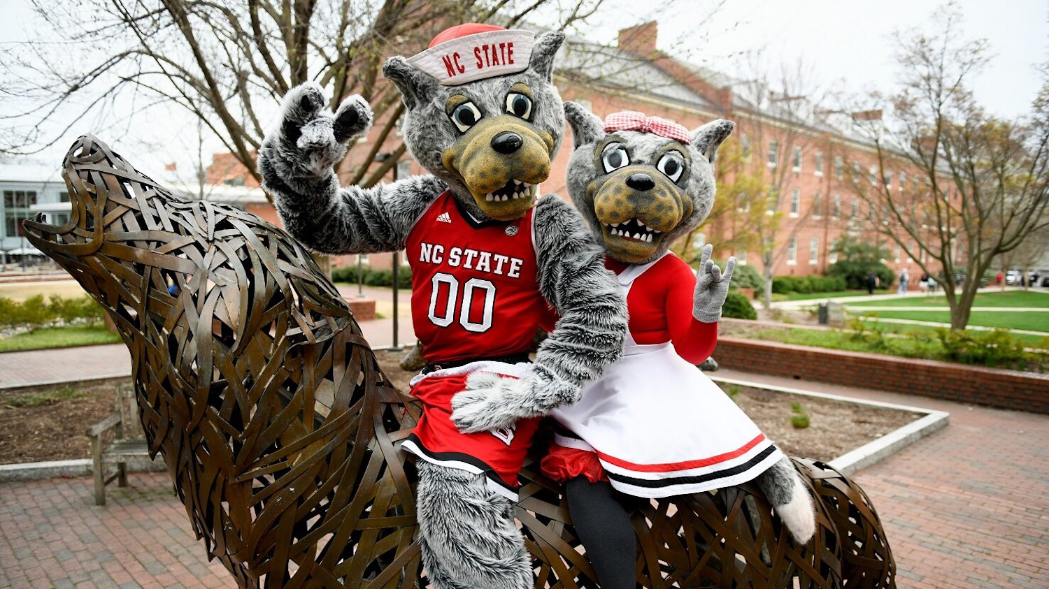 Mr. and Ms. Wuf sitting on a wolf statue in Wolf Plaza while holding up the wolfie hand sign.