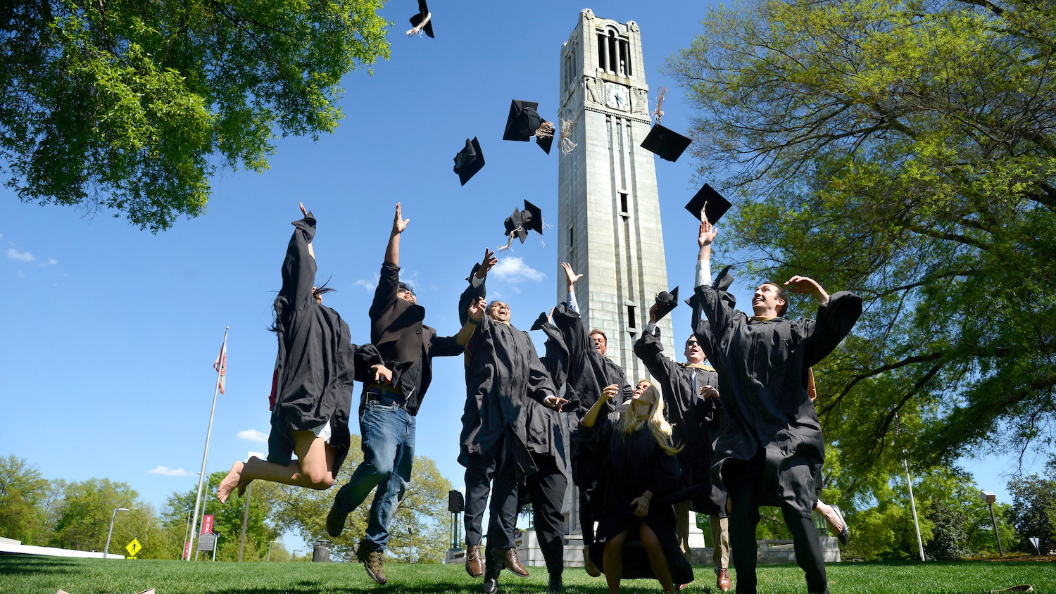 NC State graduate students in their graduation gowns throwing their caps into the air and jumping in front of the Memorial Belltower at Henry Square.
